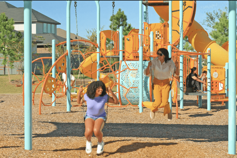 Two women smiling on swings at a colorful playground with orange play structures and slides.