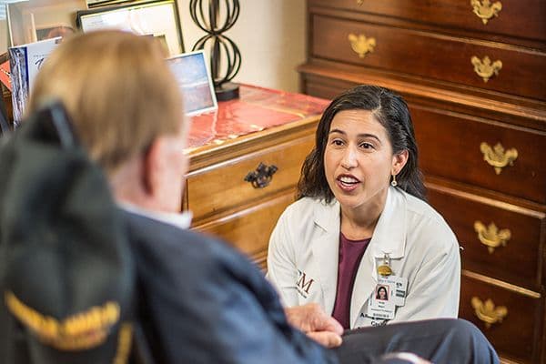 Doctor in white coat with ID badge speaks with a seated patient in an office with wooden furniture.