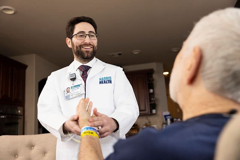 Smiling Harris Health doctor in white coat holds the hand of an elderly patient during a home visit.