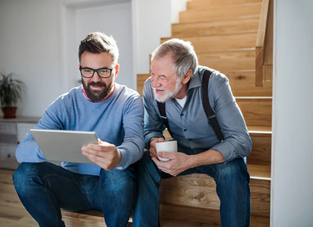 A younger man shows a tablet to a smiling older man holding a mug as they sit on wooden stairs.
