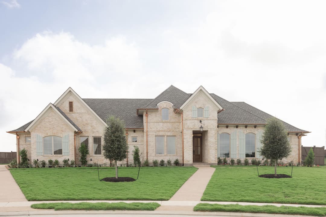 Large single-story cream brick house with gray gabled roof, blue shutters, manicured lawn and young trees.