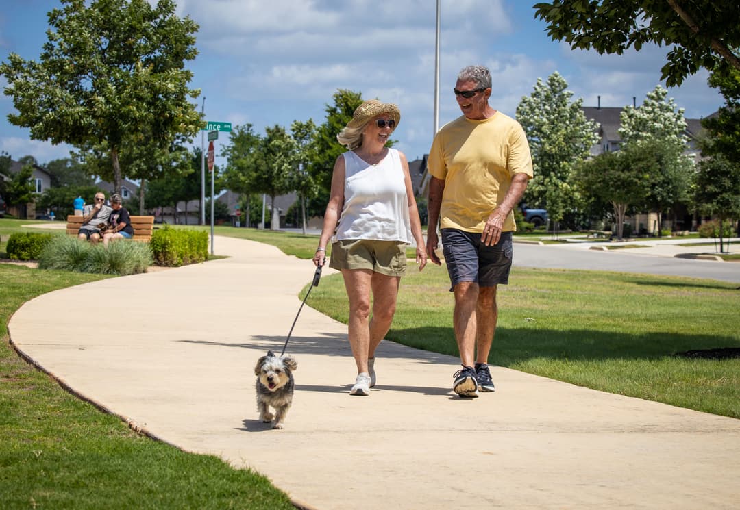 Smiling older couple walking a small gray dog on a sunny neighborhood sidewalk.