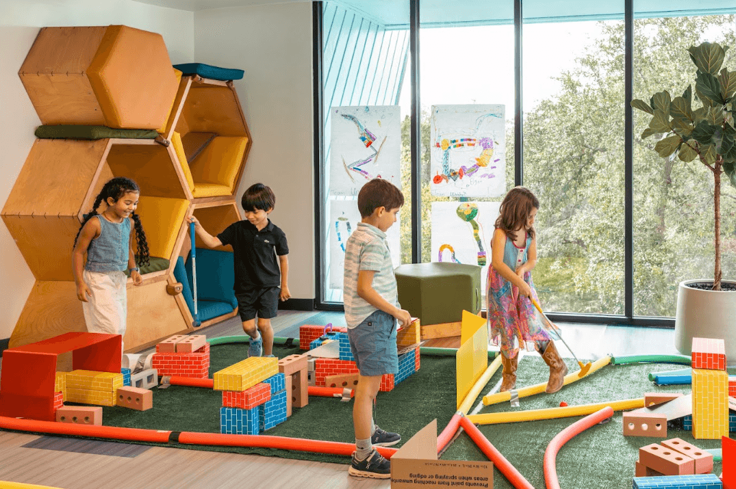 Four children play with colorful foam blocks and pool noodles in a bright playroom with hexagonal wooden shelves.