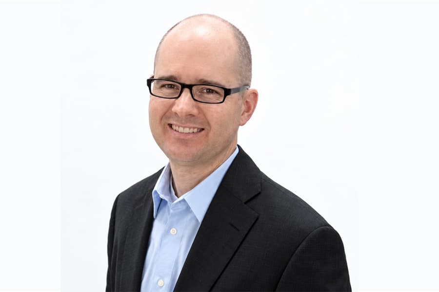 Jason Casity with glasses wearing a dark blazer and light blue shirt against a white background.