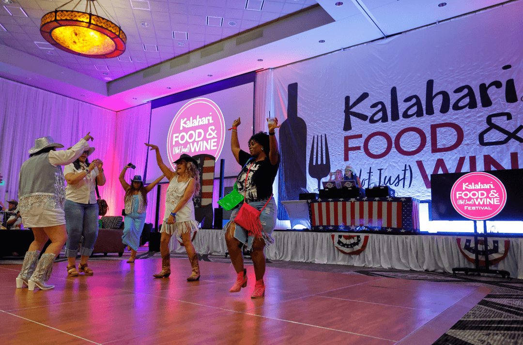 Women in cowboy boots and hats dance on a floor at the Kalahari Food & Wine Festival near a DJ booth.