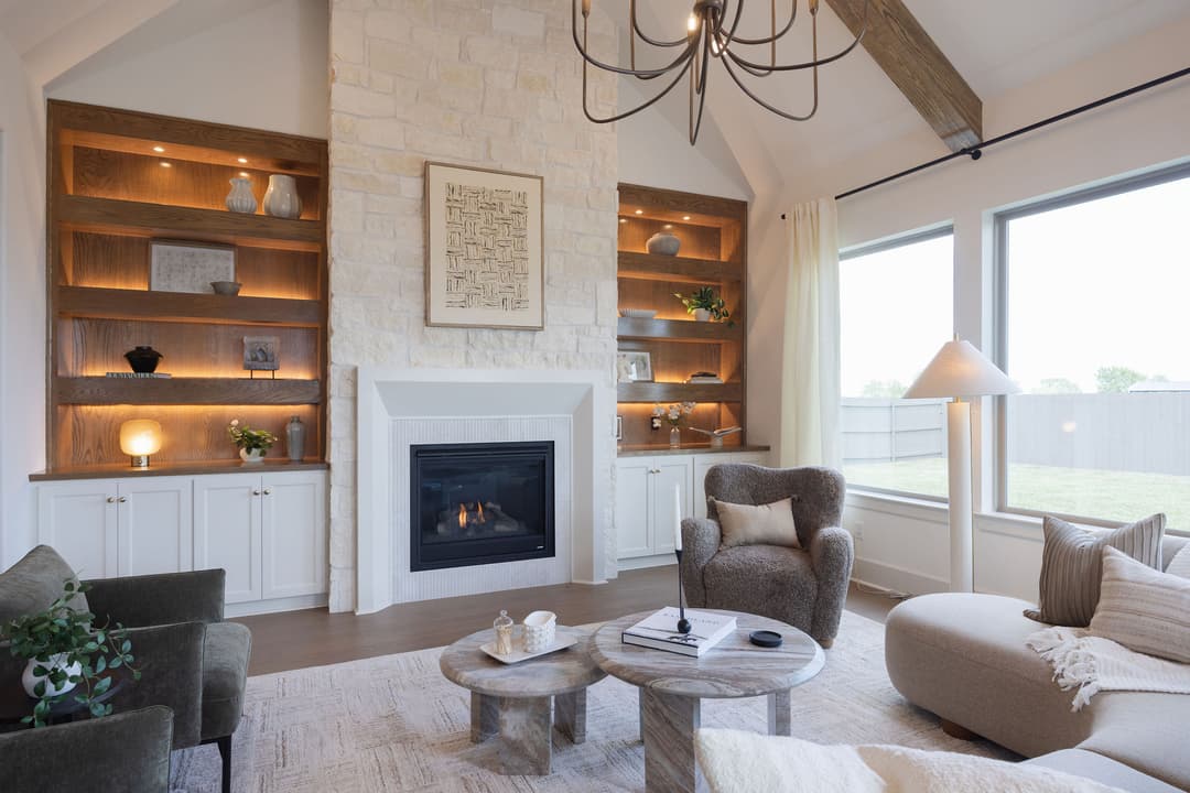 Modern living room with stone fireplace, lit wood shelves, vaulted beamed ceiling, and cozy seating by large windows.