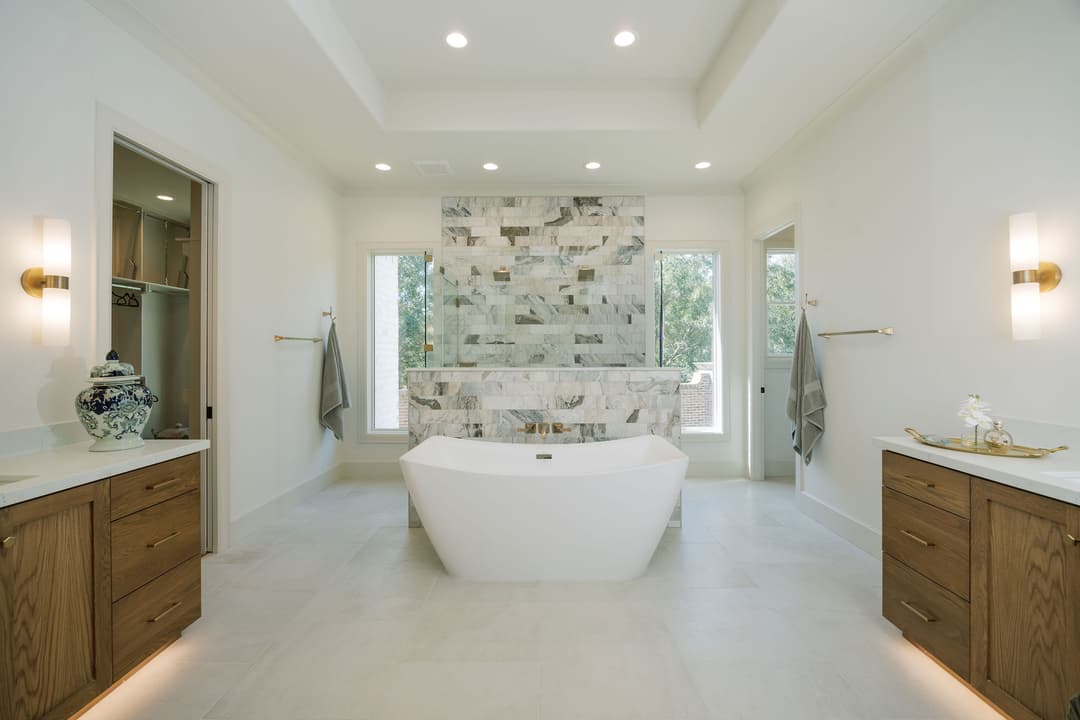 Luxury bathroom with freestanding tub, marble tile wall, twin wood vanities, and glass shower between windows.