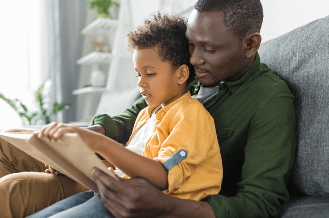 Father and young son read a book together while sitting on a couch at home.