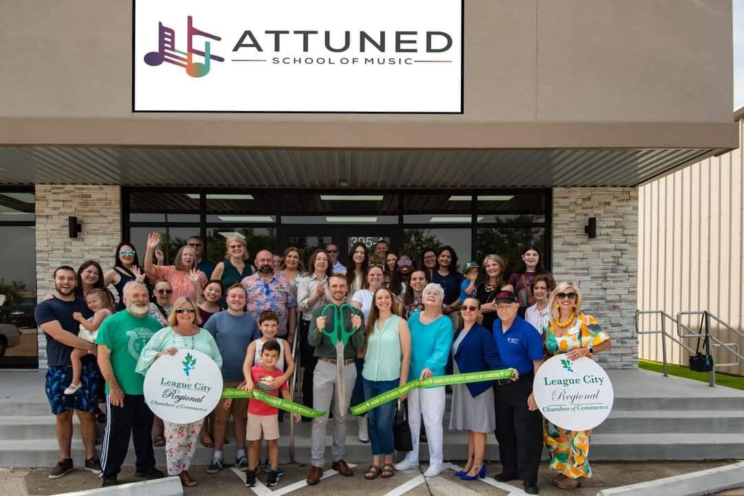 Group at ribbon-cutting ceremony outside Attuned School of Music with League City Regional Chamber of Commerce signs.