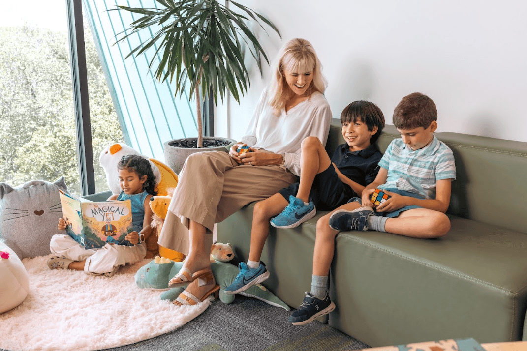 Woman on green sofa with two boys holding puzzle cubes; young child reads a book on the floor nearby.