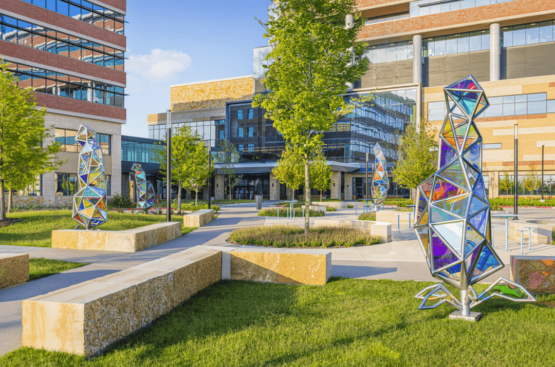 Colorful geometric glass sculptures in a landscaped courtyard among trees and modern office buildings.