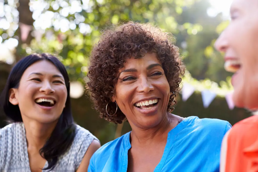Three women laughing together outdoors at a garden gathering with bunting flags in the background.