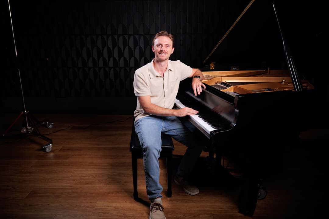 Smiling man in beige shirt and jeans sits at a grand piano in a dark studio.