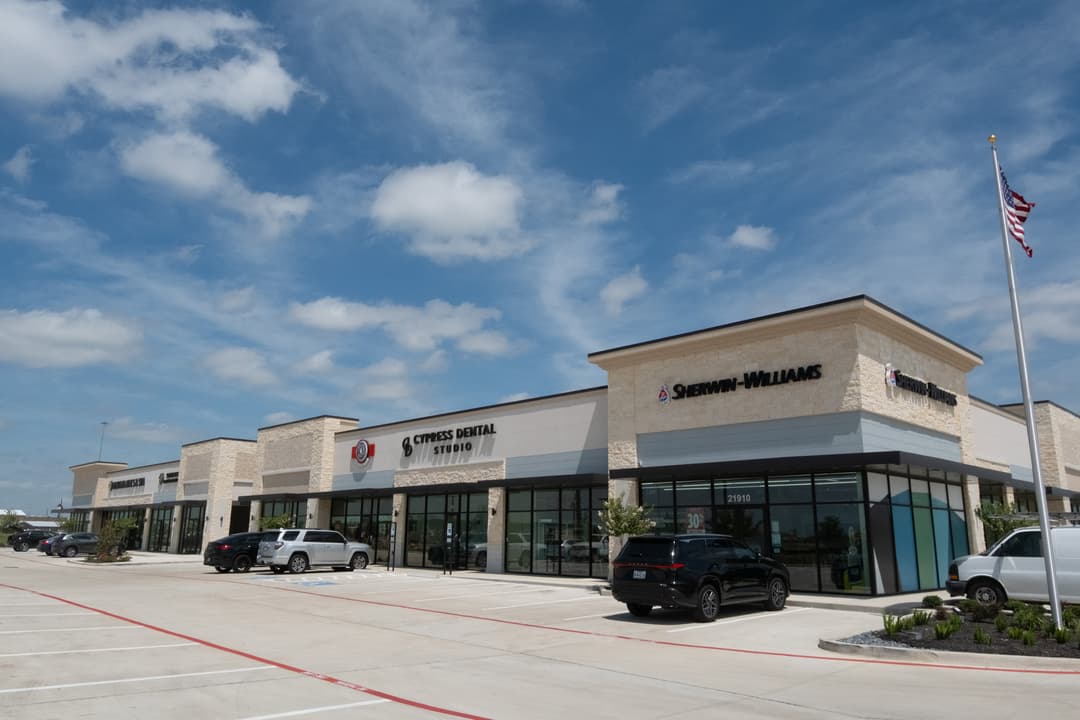 Strip mall with Sherwin-Williams and Cypress Dental Studio, parked cars, and US flag under blue sky.