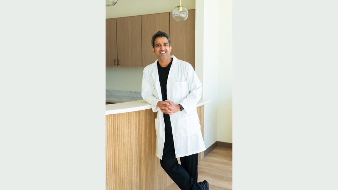 Smiling man in white lab coat leans against a reception counter in a modern medical office.