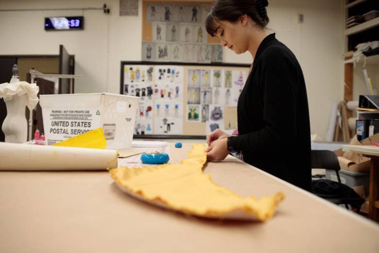 Woman in black works with yellow fabric at a table in a costume design studio with sketches on the wall.