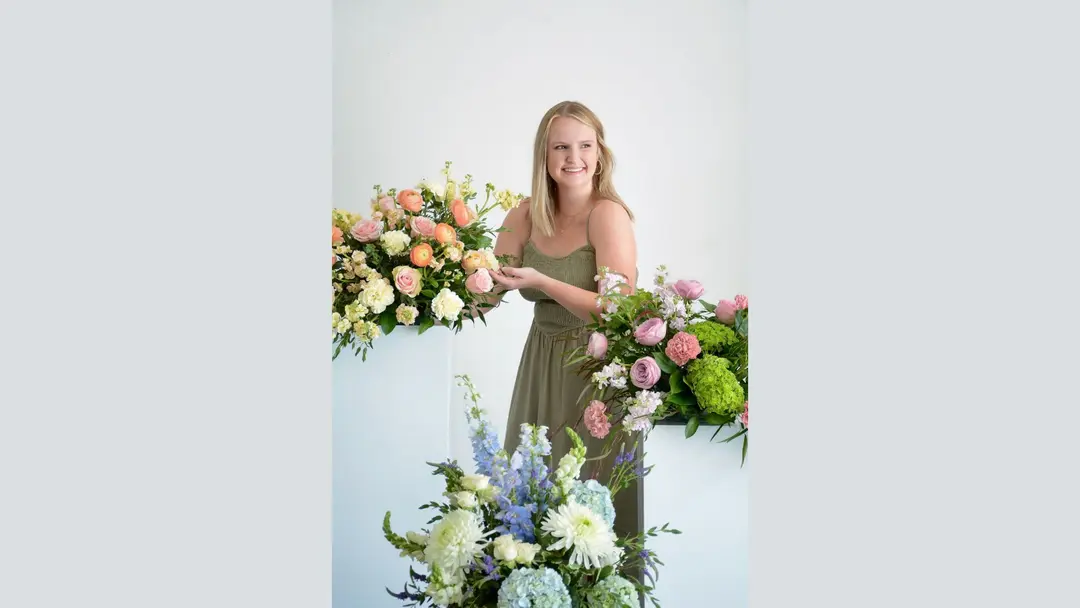Smiling woman in green dress arranges colorful floral displays with roses, hydrangeas and delphiniums.