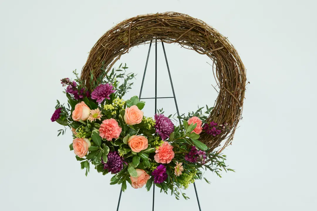 A straw wreath with pink roses and purple flowers against a gray background