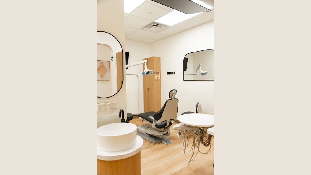Modern dental exam room with reclining chair, overhead light, wood cabinet, sink, and instrument tray.