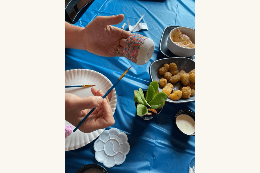Hands holding a painted pot and brush at a table with fried snacks, dip, and a succulent.