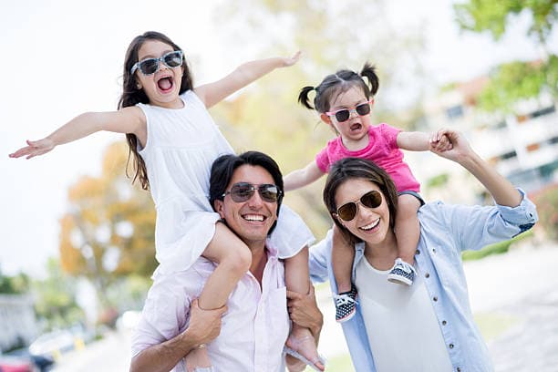 Smiling parents in sunglasses carry two young girls on their shoulders outdoors, all wearing sunglasses.