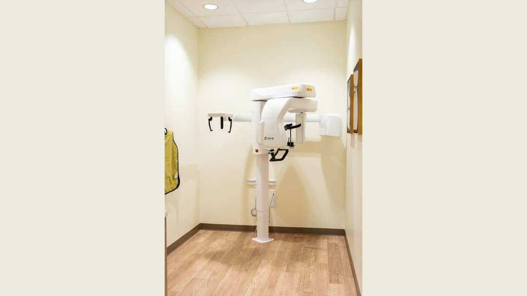 Dexis panoramic dental X-ray machine in a small room with a yellow lead apron on the wall.