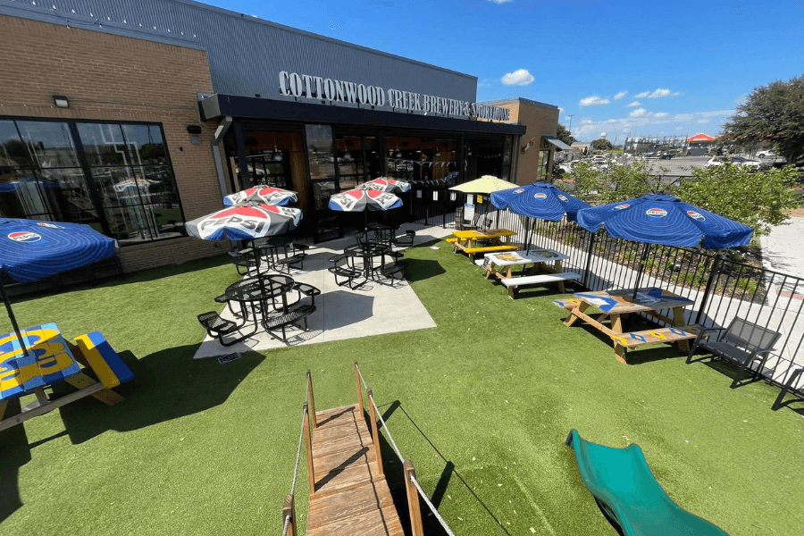 Outdoor patio at Cottonwood Creek Brewery with Pepsi umbrellas, picnic tables, and a playground on artificial turf.