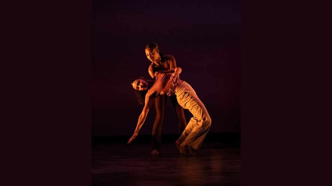Two dancers perform on a dimly lit stage, one arching backward while the other supports them.