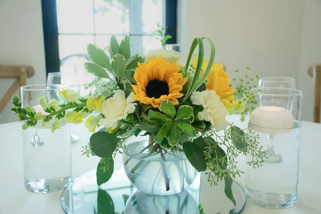 Floral centerpiece with sunflowers and greenery in glass vase, flanked by floating candles on a table.