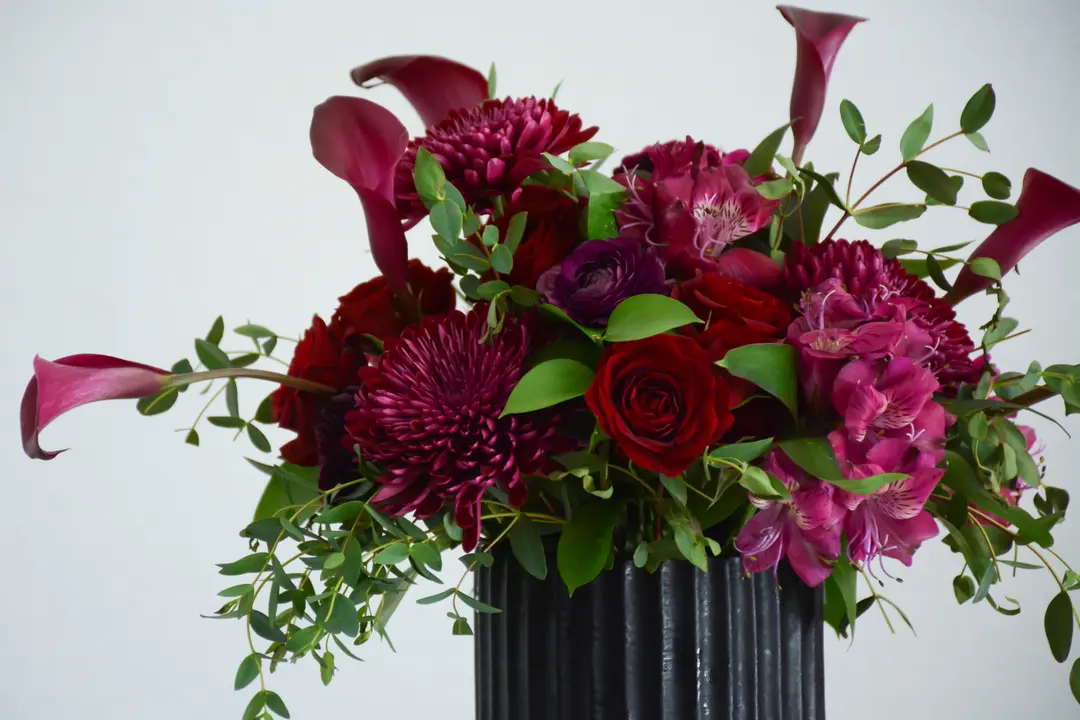 Arrangement of dark purple flowers and green leaves in a black vase, set in front of a gray background