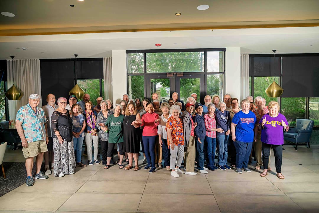 Large group of smiling seniors posing together in a bright lobby with tall windows.