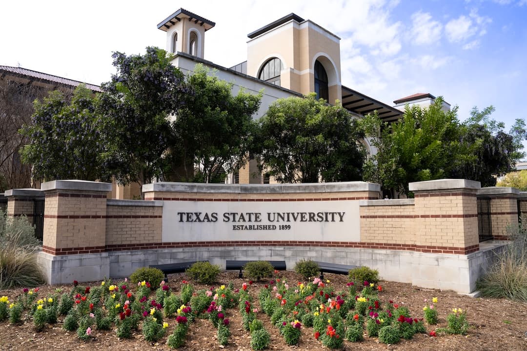 Texas State University entrance sign reading "Established 1899" with flowers, trees and a campus building behind.