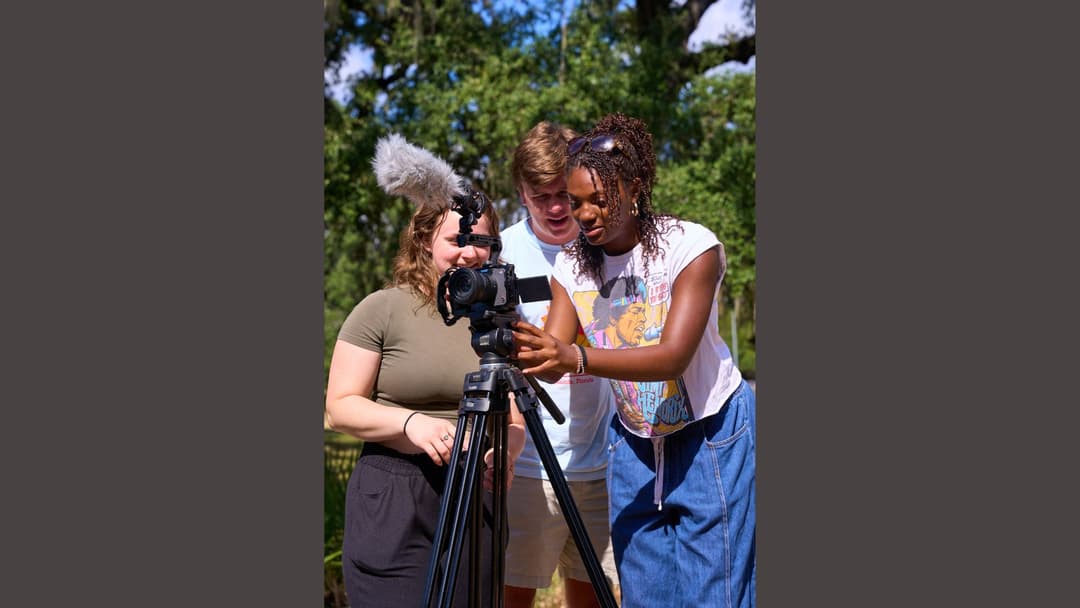 Three young people gather around a tripod-mounted camera with a microphone, filming outdoors among trees.