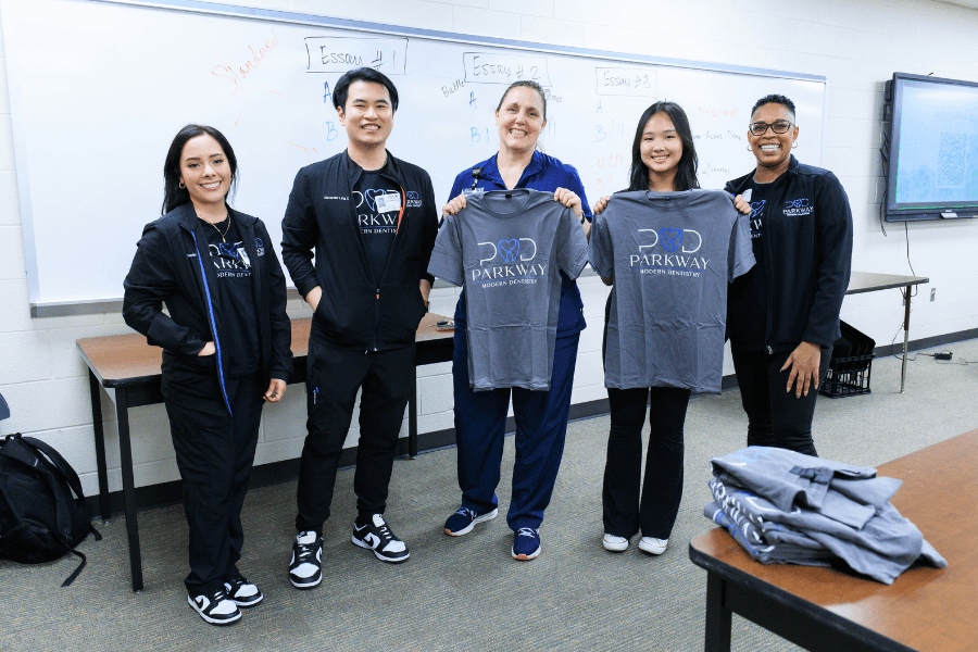 Five people smiling in a classroom, two holding gray Parkway Modern Dentistry T-shirts, whiteboard behind them.