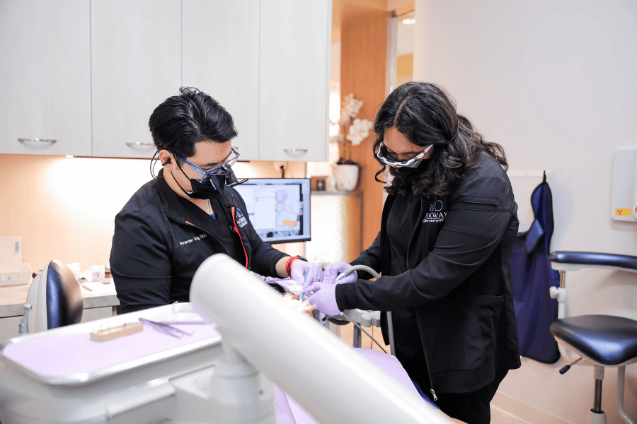 Two dental professionals in masks and gloves work together on a patient in a treatment room.