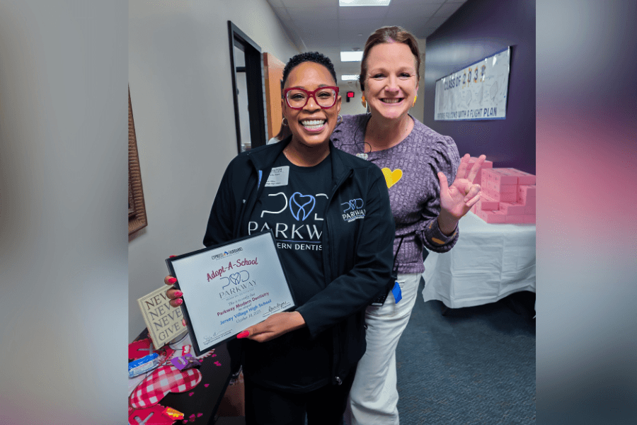Two women smiling; one holds a framed Adopt-A-School certificate from Parkway Modern Dentistry.