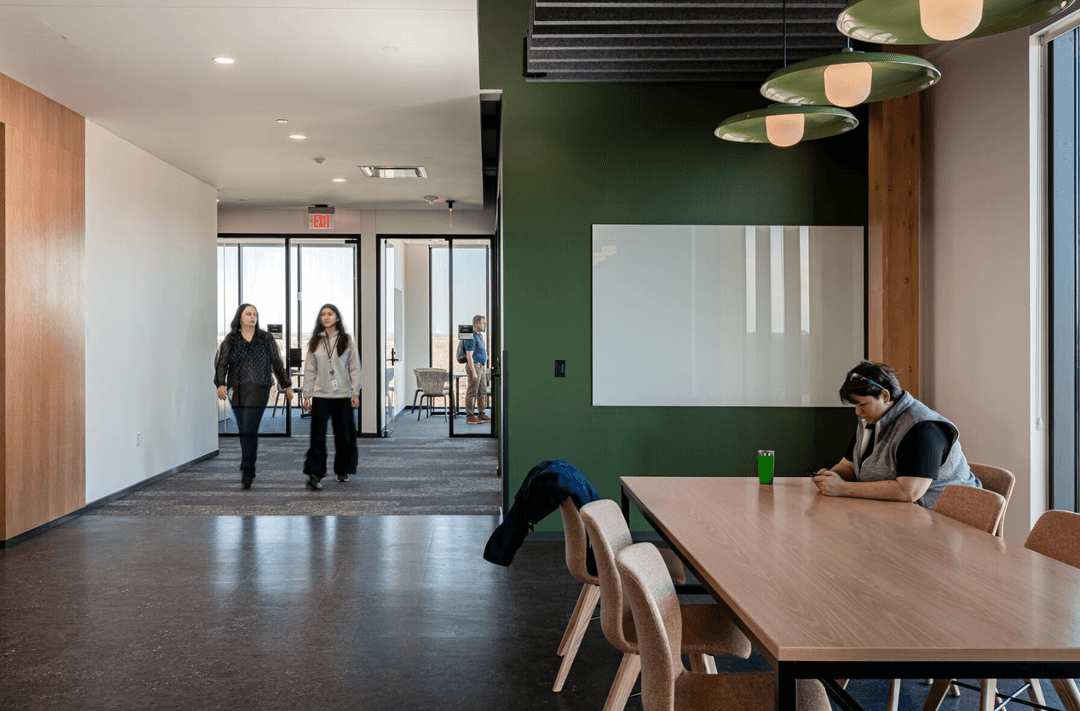 Woman seated at wooden table checks phone in modern office with green wall and whiteboard; people walk in hallway.