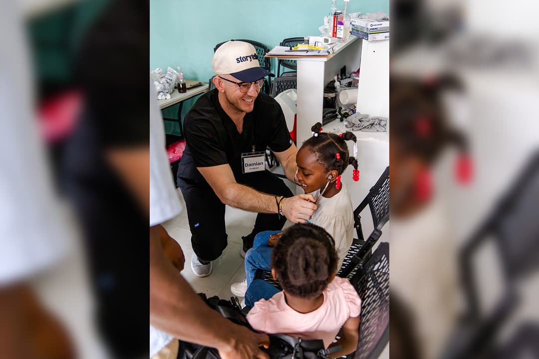 Smiling medical volunteer in scrubs lets a young girl listen through his stethoscope at a clinic.