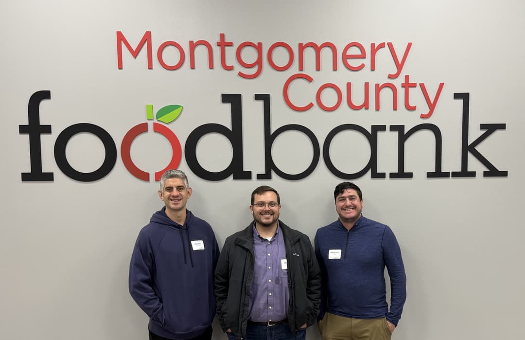 Three men wearing name tags smile in front of the Montgomery County Food Bank logo on a wall.