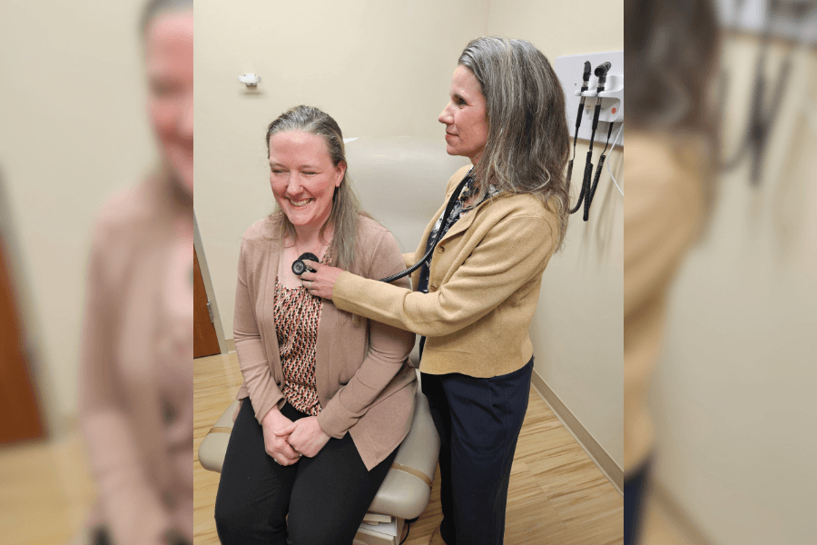 A medical provider uses a stethoscope to listen to a smiling seated patient's chest in an exam room.
