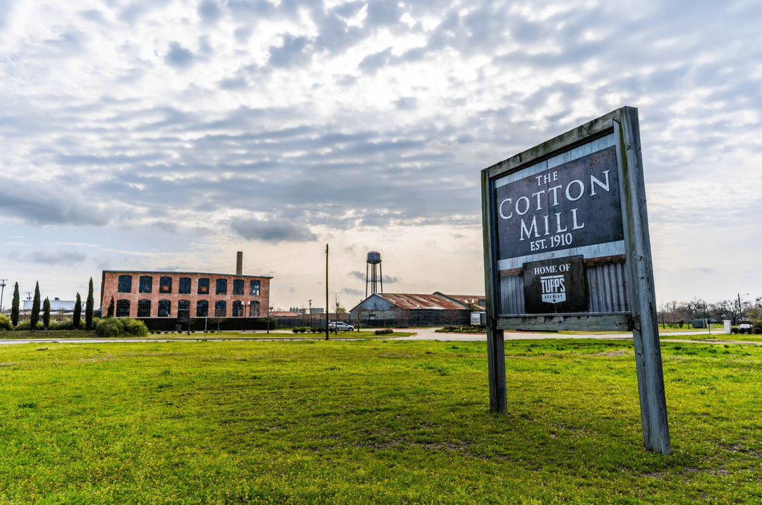 Sign reading "The Cotton Mill Est. 1910, Home of Tupps Brewery" with brick mill building and water tower behind.
