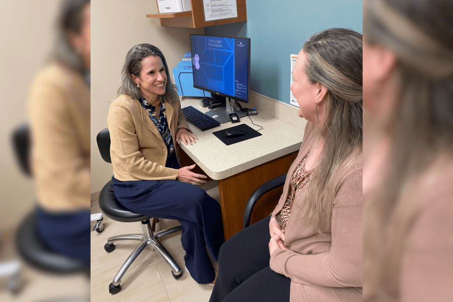 Smiling healthcare provider on a stool talks with a patient in an exam room with a computer workstation.