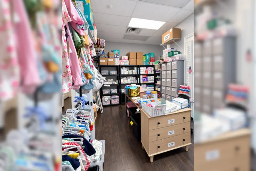 Baby supply room with racks of children's clothing, shelves of diapers and boxes, and a dresser with organized bins.