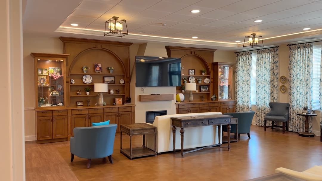 Cozy common room with wall-mounted TV, fireplace, wooden shelving, blue armchairs, sofa, and decorative built-in cabinets.