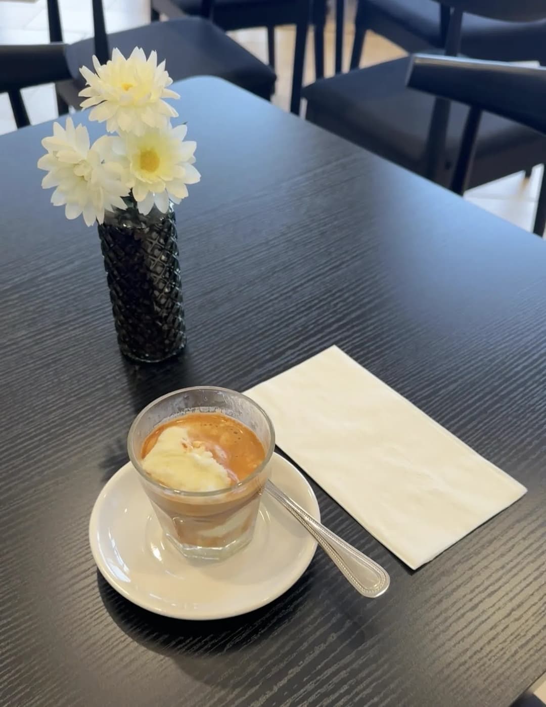 Affogato in a glass with spoon and napkin on a black café table beside a vase of white flowers.