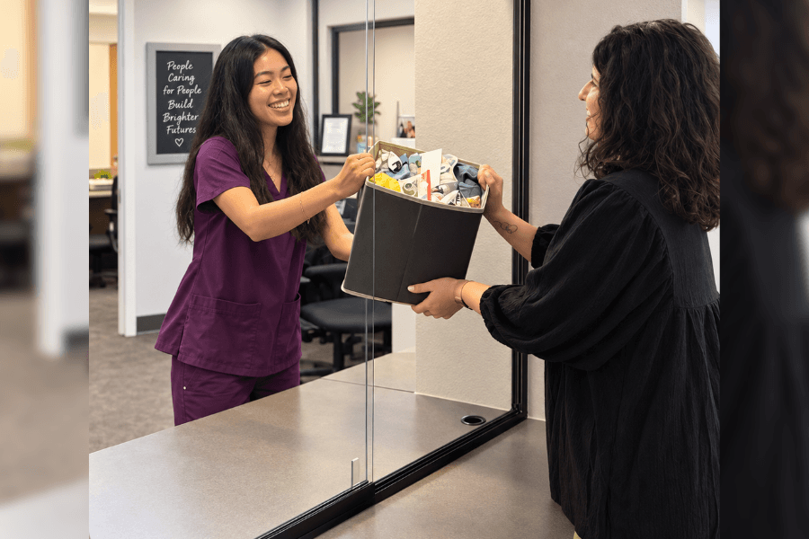 Woman in purple scrubs hands a bin of supplies to another woman across an office counter.