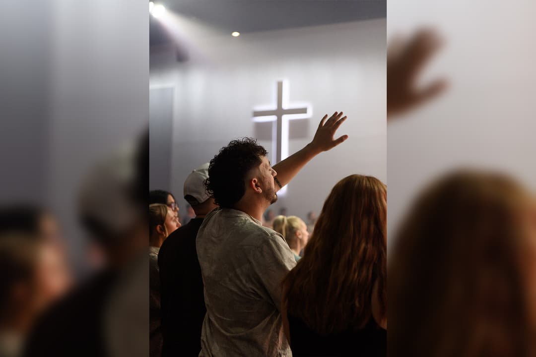 Man raises hand in worship toward an illuminated cross during a church service, surrounded by other attendees.
