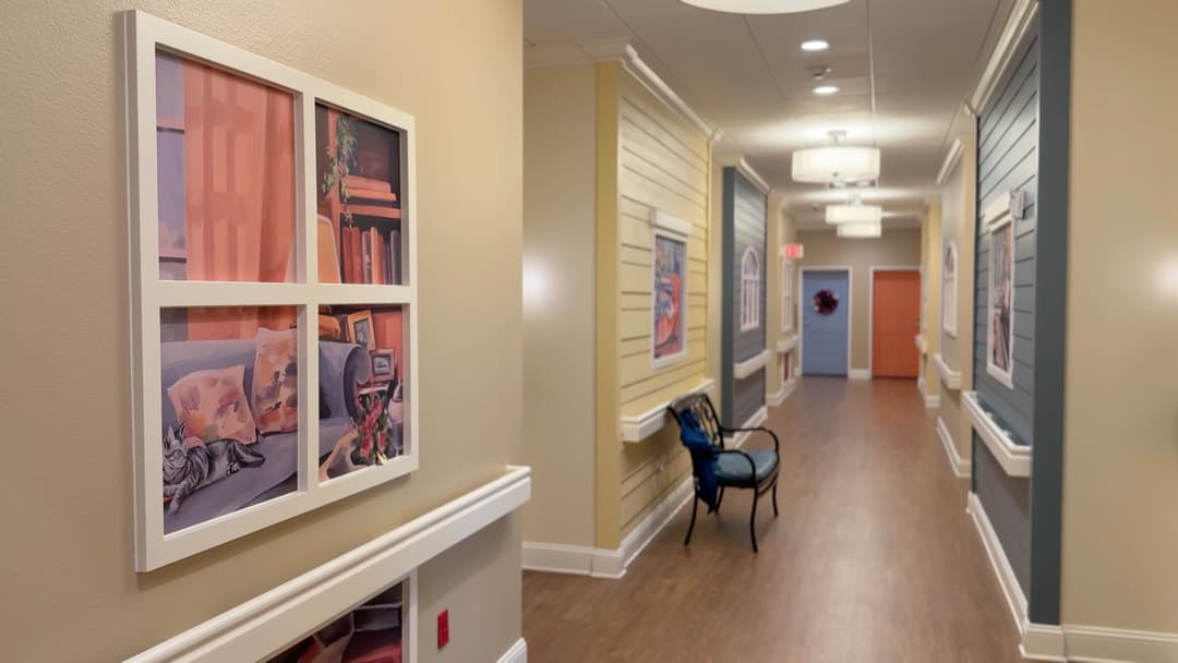 Care facility hallway with framed artwork on walls, a bench, and colorful apartment doors along the corridor.