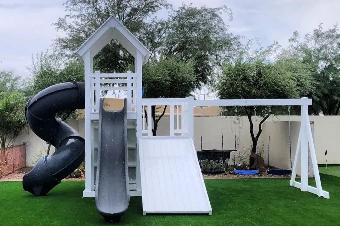 White backyard playset with gray slides, spiral tube, and swings on artificial grass near trees.