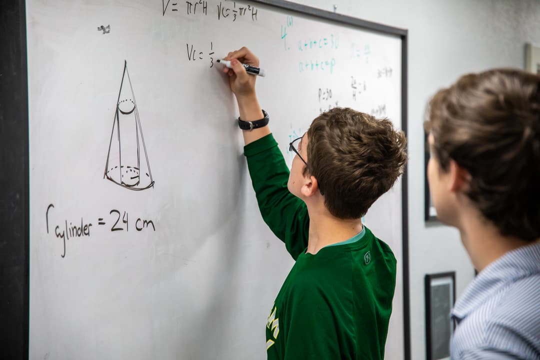 Student in green shirt writes geometry volume formulas on a whiteboard while another student watches.